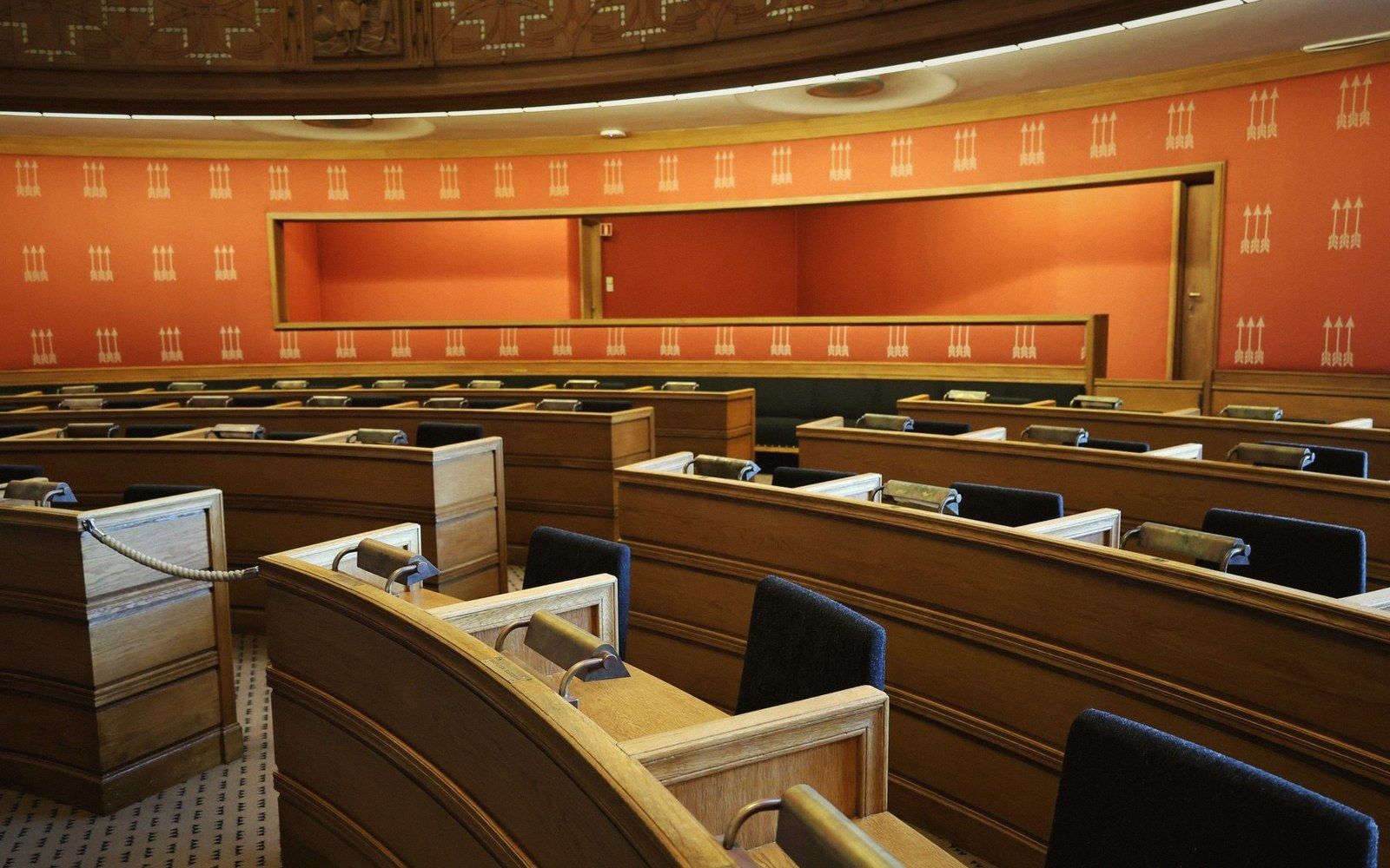 Rows of empty wooden desks and chairs in a curved assembly hall with patterned orange walls