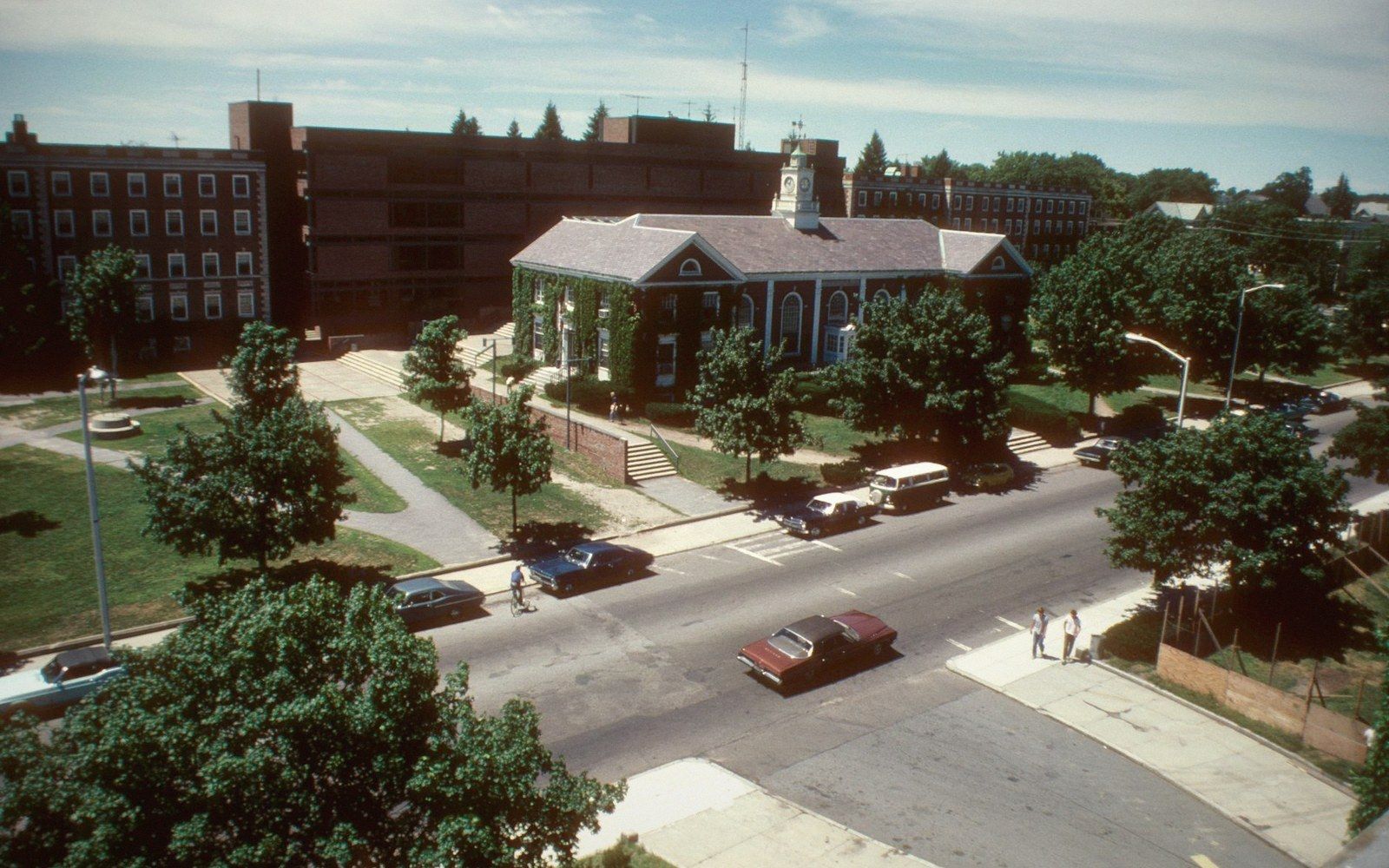 Aerial view of a clock tower building on a tree-lined campus street with parked cars